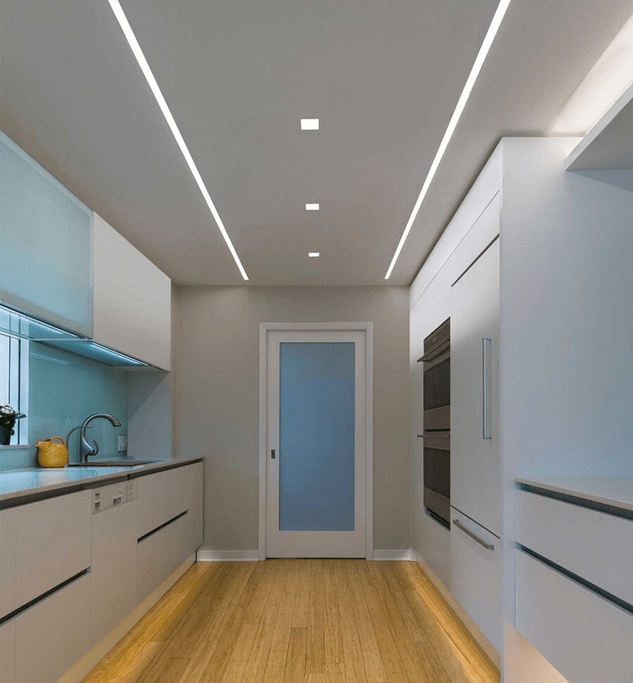 نمایی از طراحی نورپردازی لایه‌ای در آشپزخانه مدرن، با استفاده از لاین نوری توکار LED در سقف به عنوان نور محیطی، و نصب چراغ خطی آویز برای تأمین نور وظیفه‌ای متمرکز و دکوراتیو بر روی جزیره مرکزی.