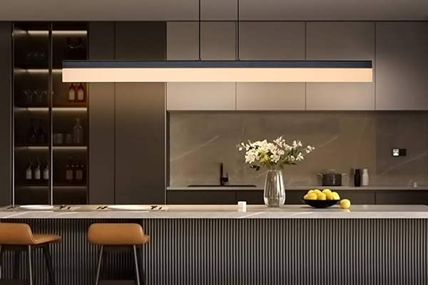 نمایی از طراحی نورپردازی لایه‌ای در آشپزخانه مدرن، با استفاده از لاین نوری توکار LED در سقف به عنوان نور محیطی، و نصب چراغ خطی آویز برای تأمین نور وظیفه‌ای متمرکز و دکوراتیو بر روی جزیره مرکزی.